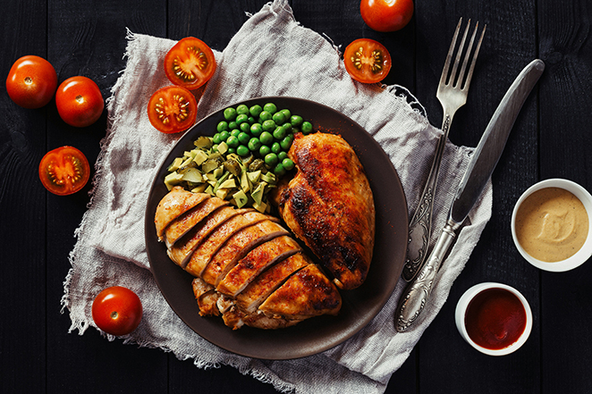 Overhead view of roasted chicken breast with green peas and diced green vegetables on a dark plate, with cherry tomatoes and small sauce dishes on a wood table
