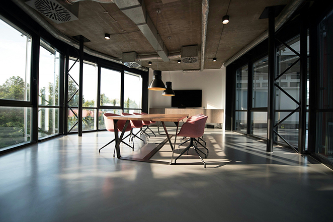 Sunlit conference room with a long wooden table, salmon-upholstered chairs, exposed concrete ceiling, and floor-to-ceiling windows, empty chairs where hybrid and return-to-office policies actually show up.