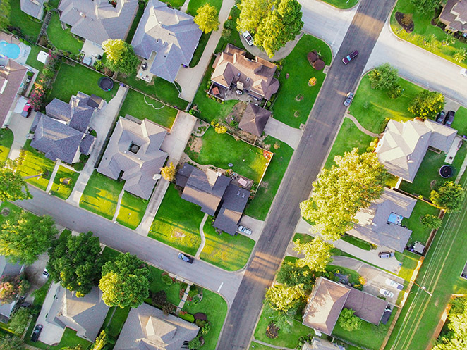 High-angle aerial photo of a suburban block on a clear day: single-family homes with shingled roofs, deep green lawns, scattered shade trees casting long shadows, two asphalt streets meeting at an intersection, and a few cars in driveways and along the curb.