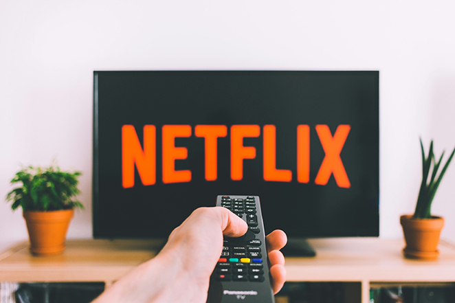First-person view: a hand holding a TV remote aimed at a flat-screen showing the Netflix logo, on a light wood stand with small potted plants—a familiar slice of the monthly streaming bill