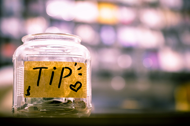 Clear glass tip jar with a hand-written "Tip" label and a small heart, folded dollar bills inside, on a dark counter with soft warm bokeh lights suggesting a cafe or bar
