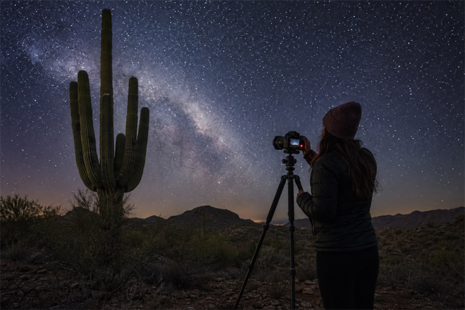 Photographer with a camera on a tripod in a desert at night, framing the Milky Way; a saguaro cactus stands to the left and the camera rear screen shows a live starry preview